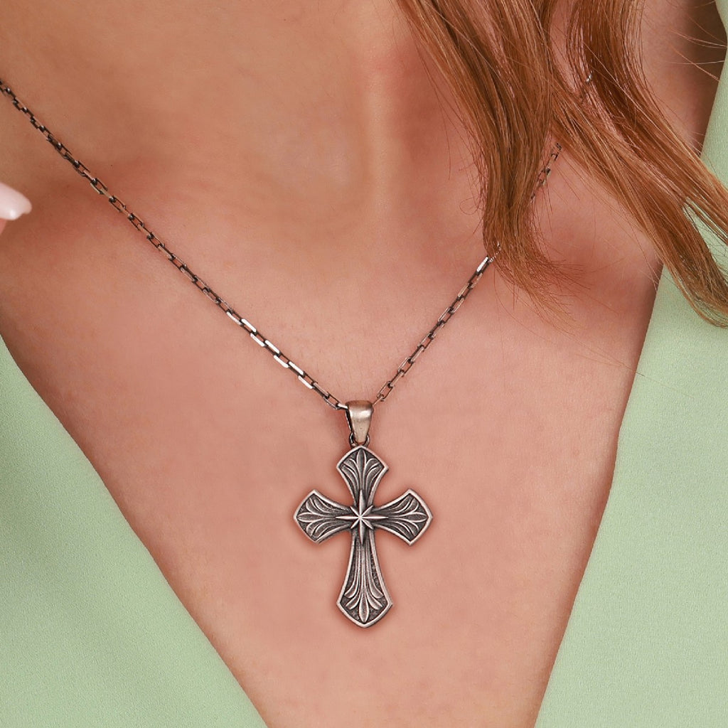 A close-up of a person wearing a sterling silver, ornate cross pendant necklace with an oxidized antique finish, set against their skin and a light green top.
