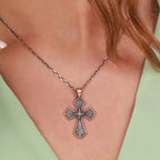 A close-up of a person wearing a sterling silver, ornate cross pendant necklace with an oxidized antique finish, set against their skin and a light green top.