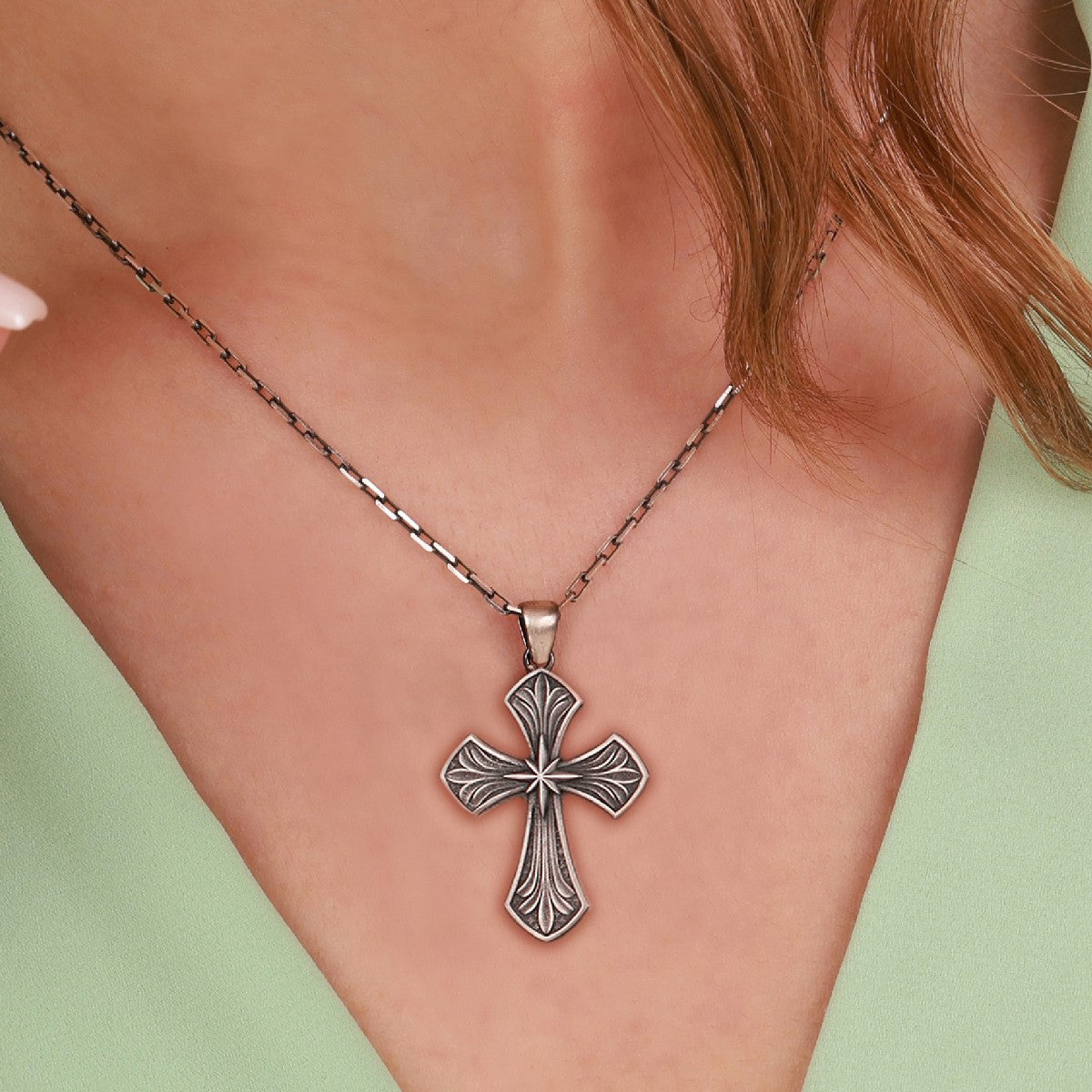 A close-up of a person wearing a sterling silver, ornate cross pendant necklace with an oxidized antique finish, set against their skin and a light green top.