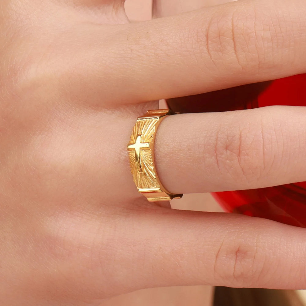 A gold ring with a cross and sunburst design, worn on a person's finger against a red background.