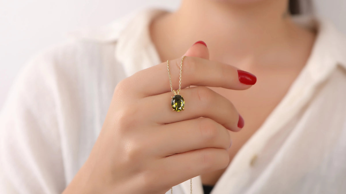 A close-up of a person's hand with red nail polish holding a delicate gold chain necklace with a small oval green gemstone pendant.