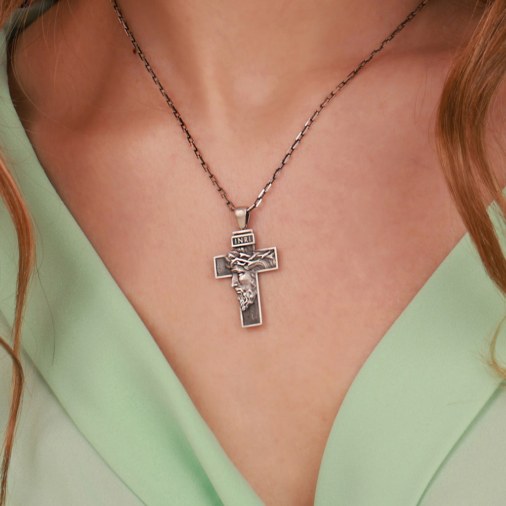 A close-up of a silver cross pendant necklace worn on a model's neck, featuring an embossed figure of Jesus Christ and the word 'INRI' on the cross, styled with a mint green top.