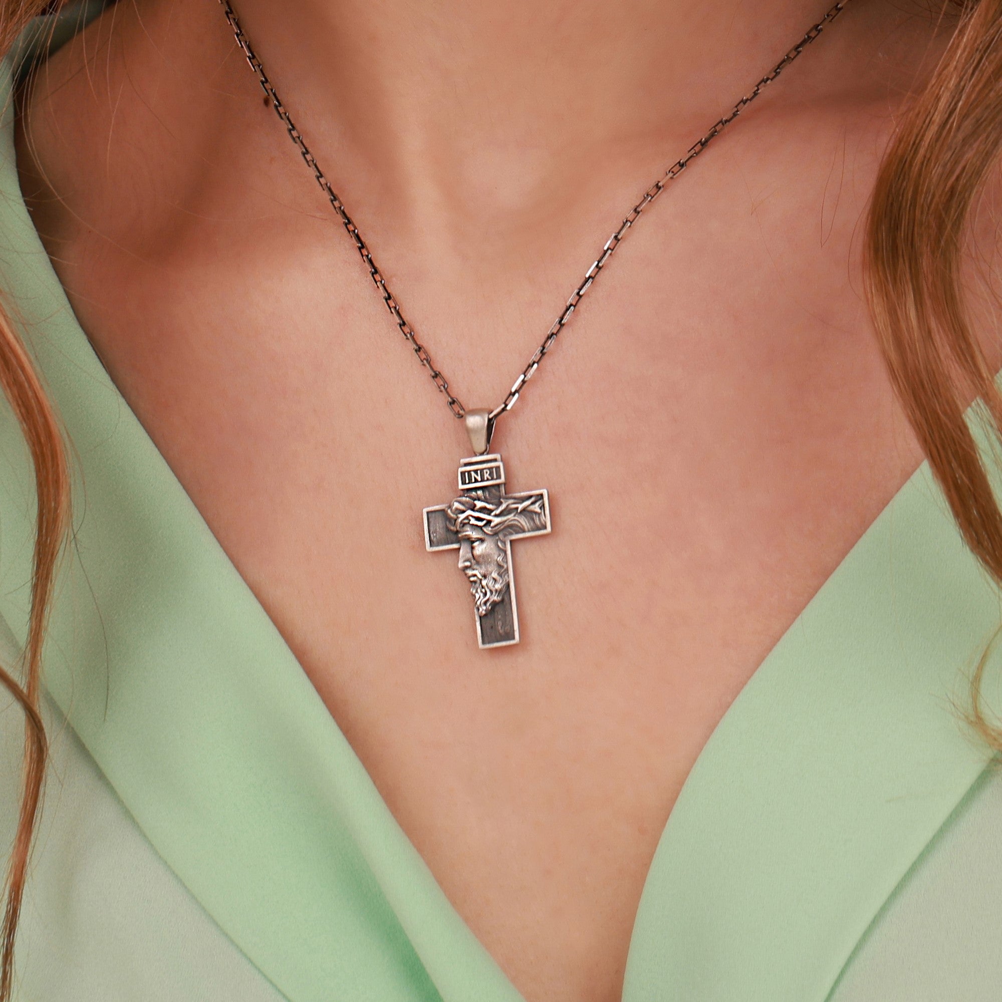 A close-up of a silver cross pendant necklace worn on a model's neck, featuring an embossed figure of Jesus Christ and the word 'INRI' on the cross, styled with a mint green top.