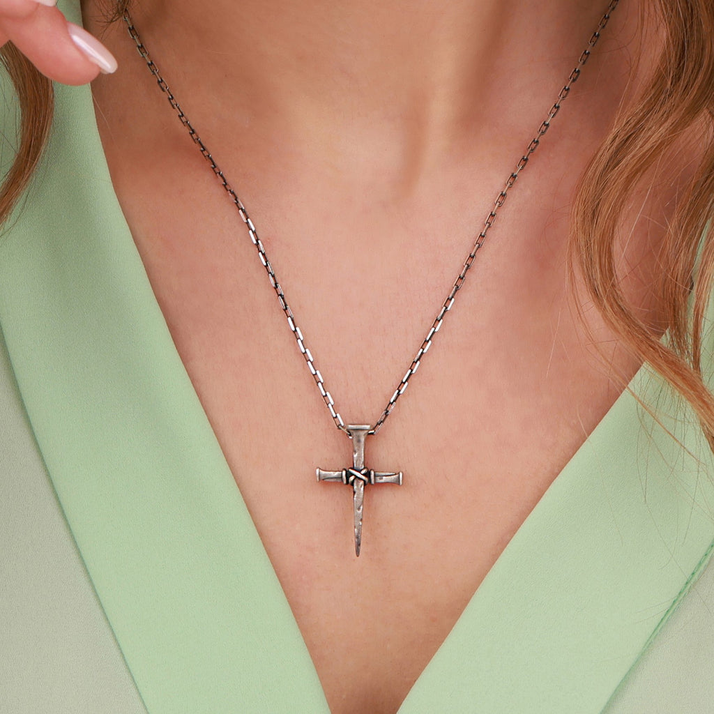 A sterling silver nail cross pendant necklace with a textured, hammered finish, worn on a model's neck over a light green V-neck top.