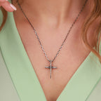 A sterling silver nail cross pendant necklace with a textured, hammered finish, worn on a model's neck over a light green V-neck top.