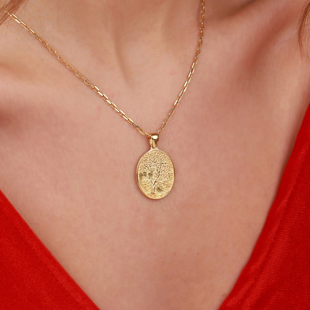 A gold oval pendant necklace with a textured tree of life design, worn on a model's neck over a red top.