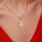 A gold oval pendant necklace with a textured tree of life design, worn on a model's neck over a red top.