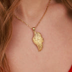 A gold angel wing-shaped pendant necklace worn on a model's neck, shown in a close-up shot with the model's skin and hair visible.