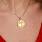 A gold necklace with a tree of life pendant featuring an engraved tree of life design, shown worn on a person's neck over a red top.