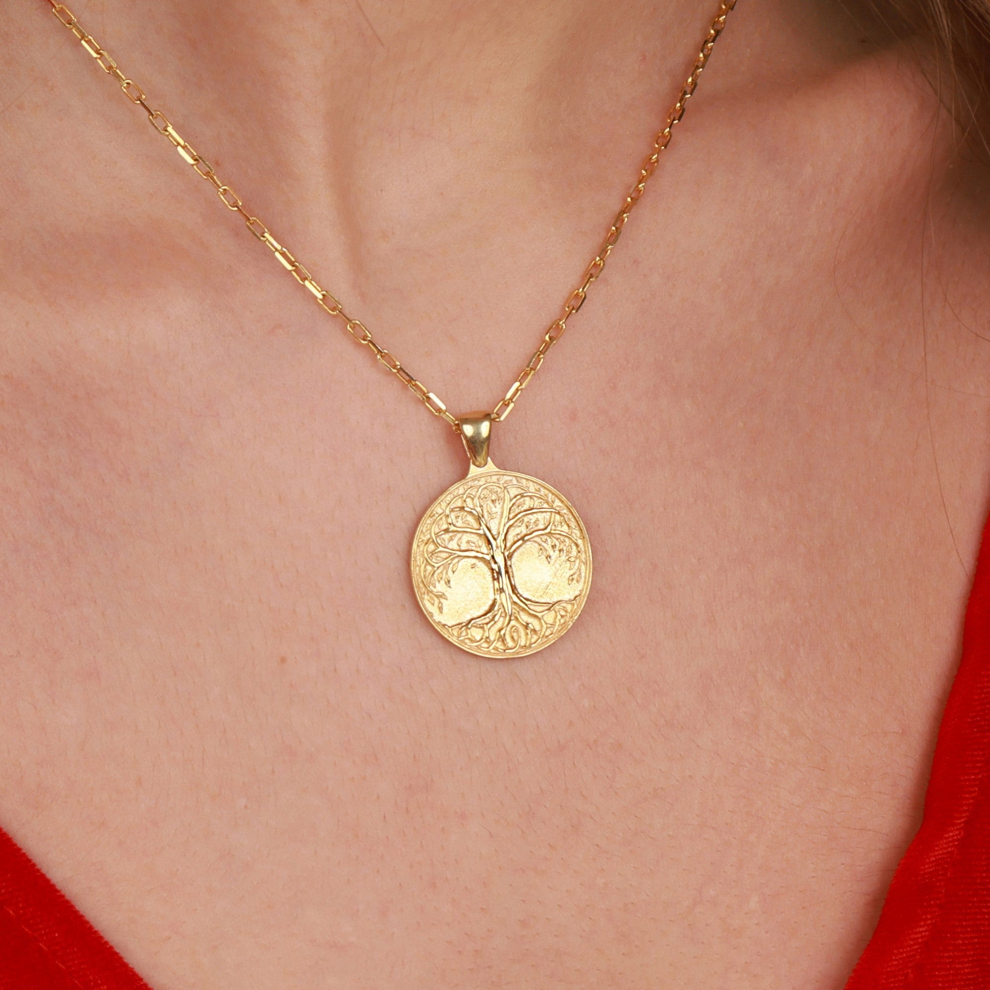 A gold necklace with a tree of life pendant featuring an engraved tree of life design, shown worn on a person's neck over a red top.