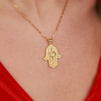 A gold Hamsa hand pendant necklace worn on a person's neck, with a red top visible at the edges.