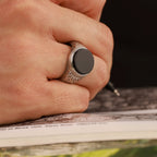 A hand wearing a silver Celtic ring with a black stone, holding a pen over a magazine.