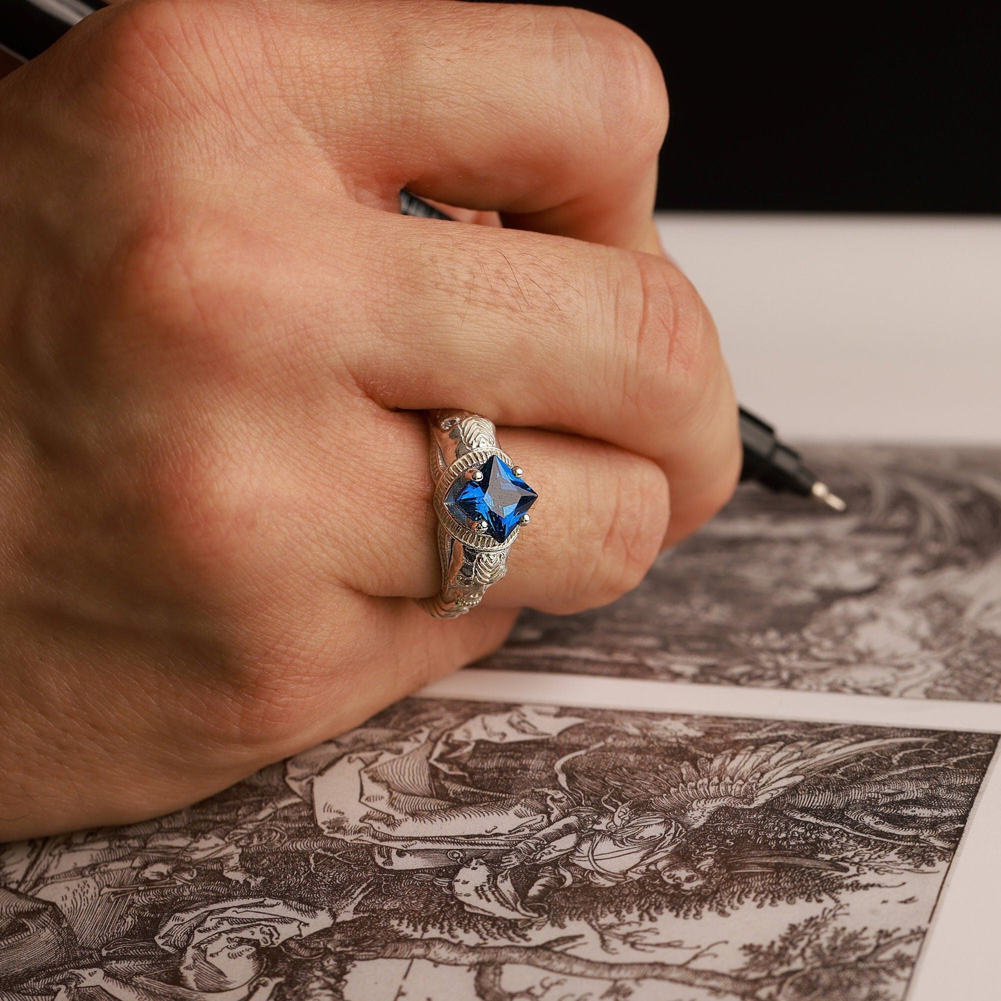 Hand with a skull ring with blue gemstone holding a pen over an engraved sheet of paper
