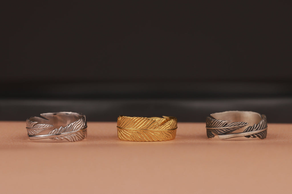 Three feather wrap rings, two silver and one gold, on a brown surface with a dark background
