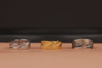 Three feather wrap rings, two silver and one gold, on a brown surface with a dark background