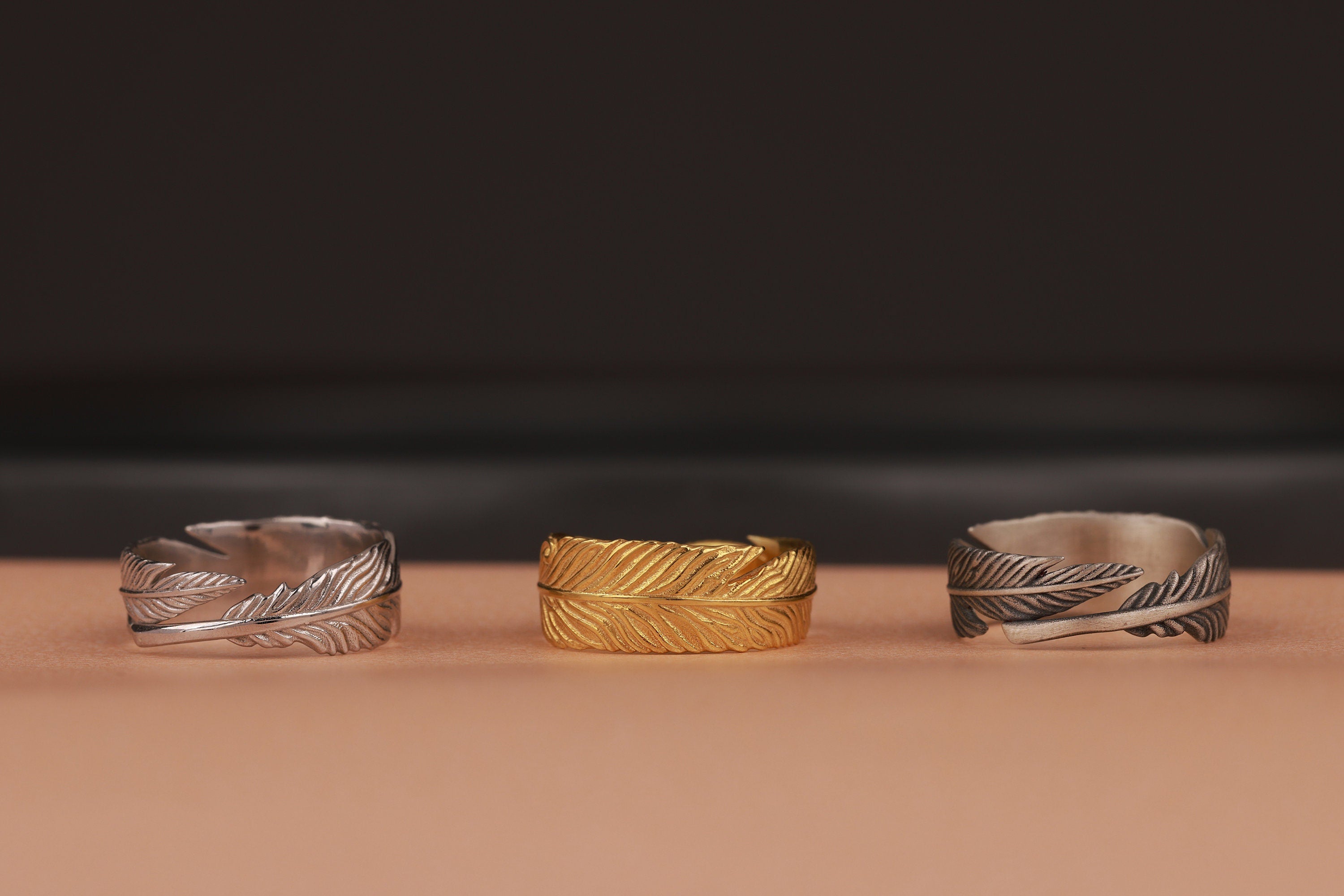 Three feather wrap rings, two silver and one gold, on a brown surface with a dark background