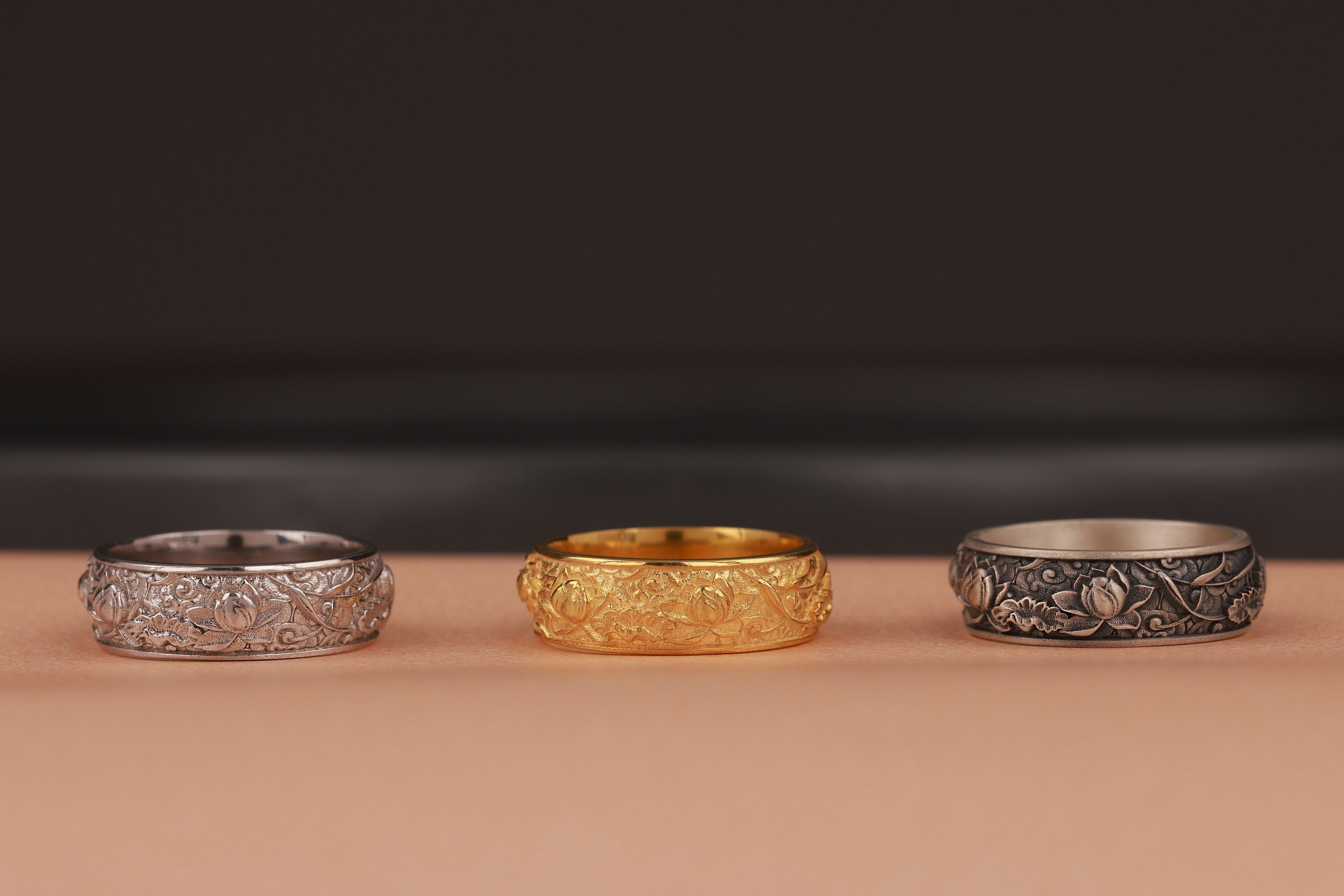 Three rings, two silver and one gold, on a beige surface with a blurred dark background