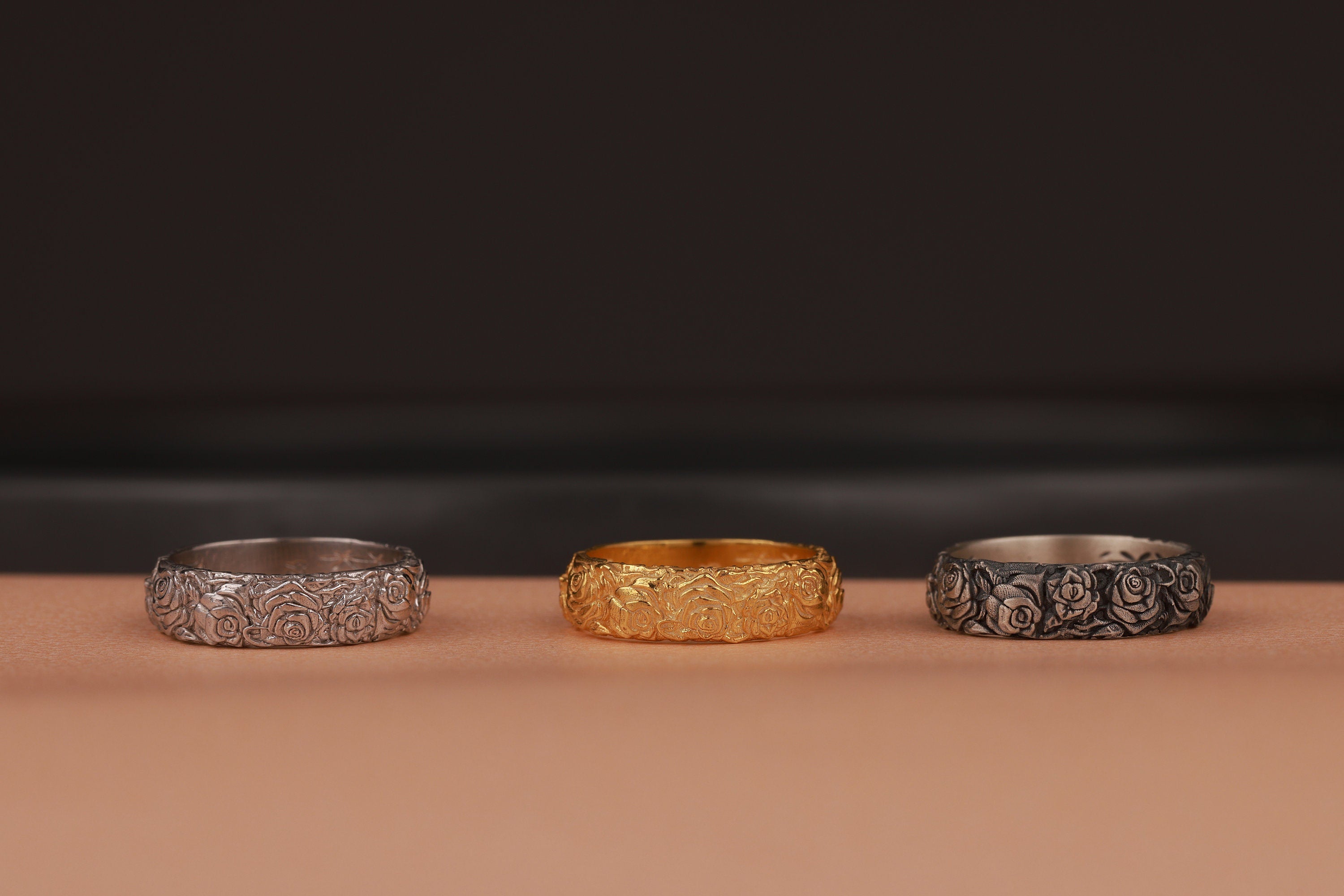 Three rose flower rings, two silver and one gold, on a brown surface with a dark background