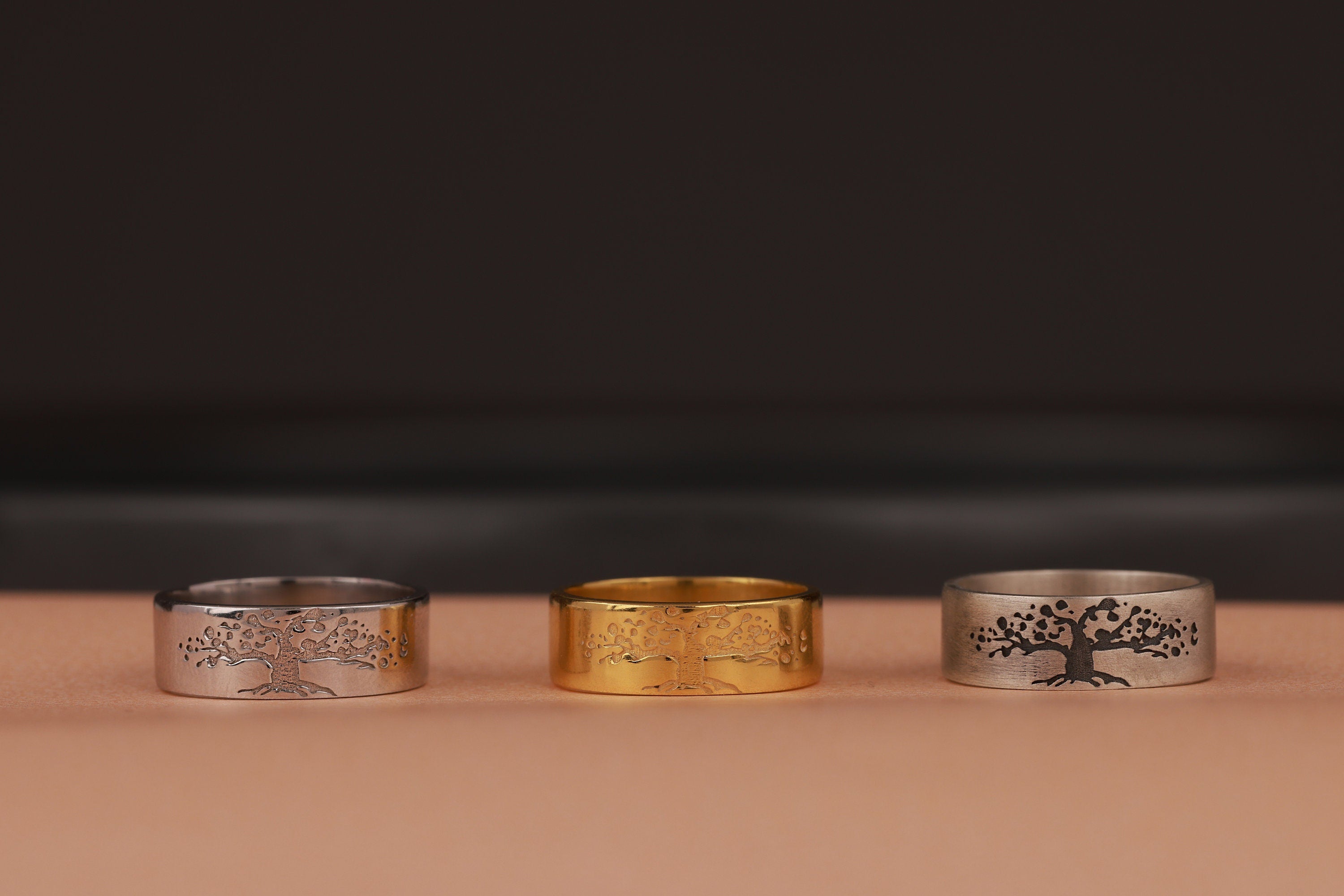 Three tree of life rings, two silver and one gold, with tree designs on a dark background