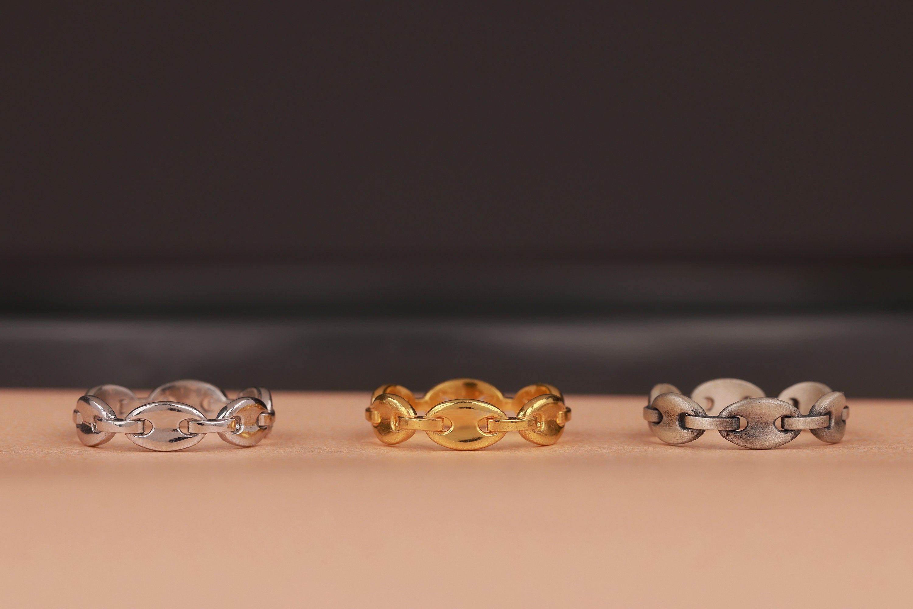 Three chain rings in silver, gold, and another silver design on a beige surface with a dark background.