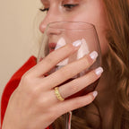 Woman holding a glass of red wine with a gold rose ring on her finger, against a beige background