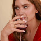 Woman in a red dress holding a glass of red wine with a gold ring against a beige background