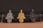 Three Hamsa hand with evil eye pendants in gold, silver, and bronze on a dark brown background.