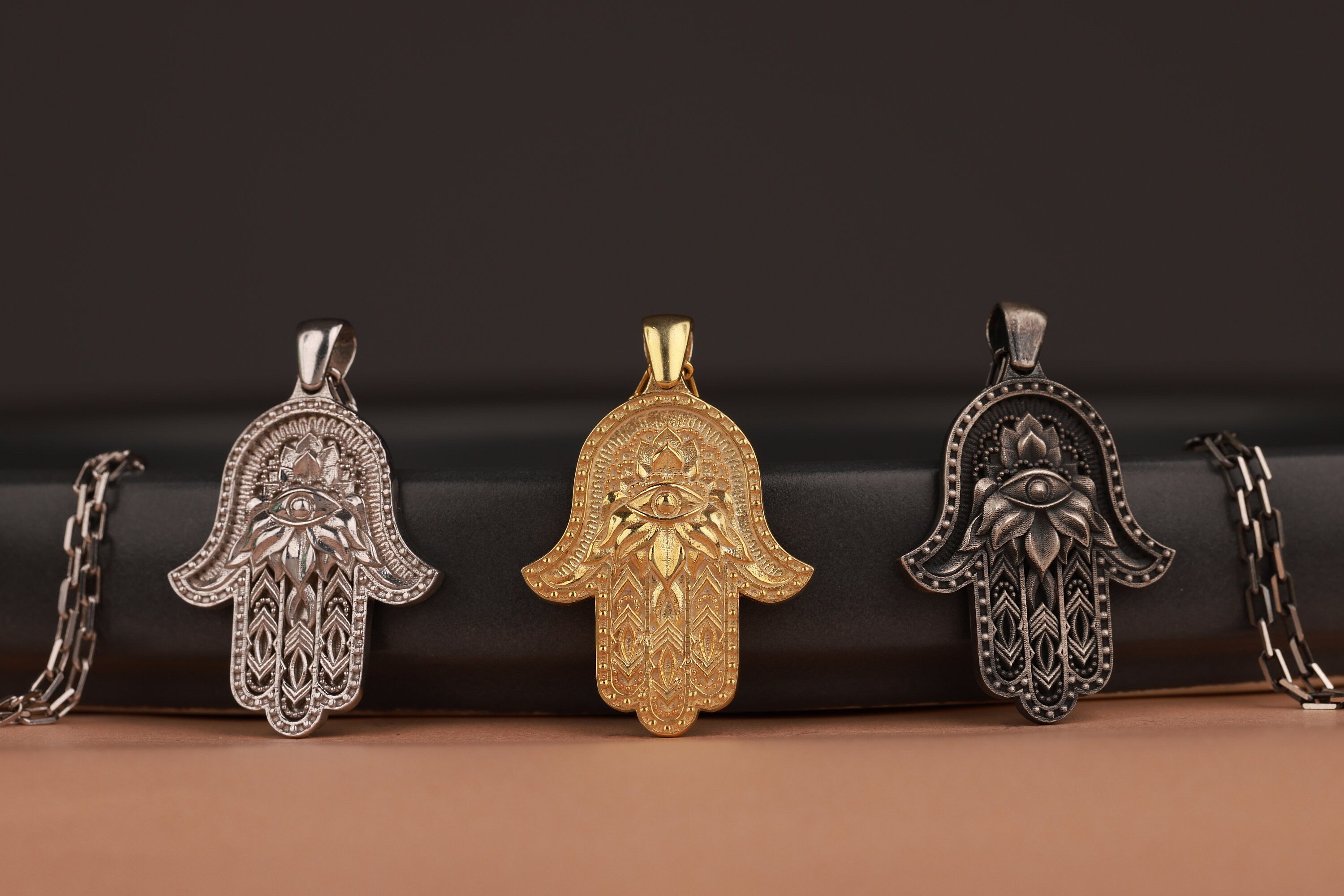 Three Hamsa hand with evil eye pendants in gold, silver, and bronze on a dark brown background.