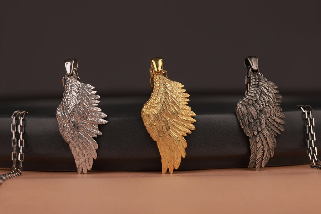 Three angel wing pendants in silver, gold, and bronze on a black and brown gradient background.