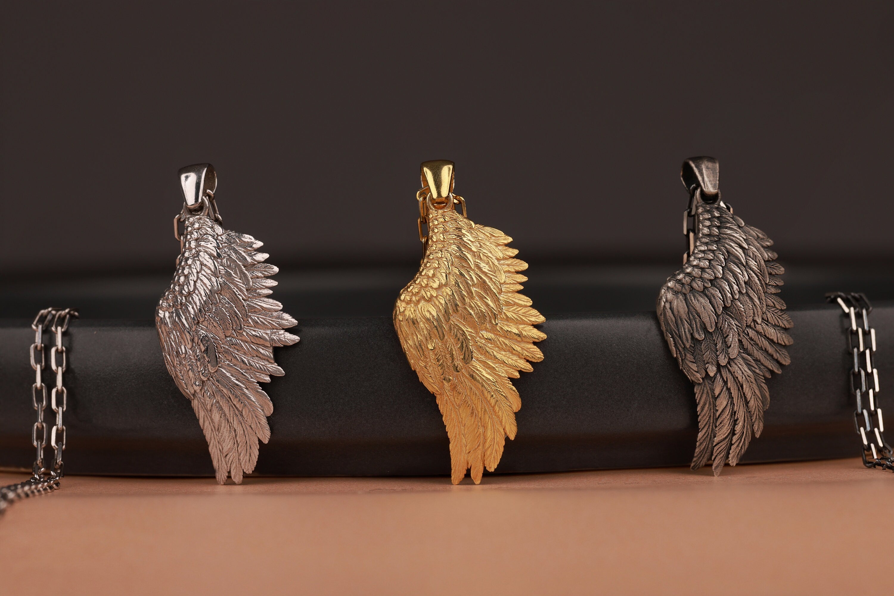 Three angel wing pendants in silver, gold, and bronze on a black and brown gradient background.