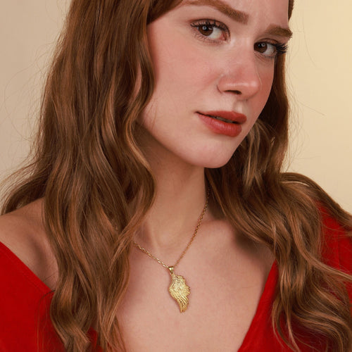 Woman wearing a gold necklace with a wing pendant against a beige background