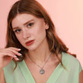 Woman wearing a green top and an oxidized necklace with a detailed tree of life pendant against a pink background