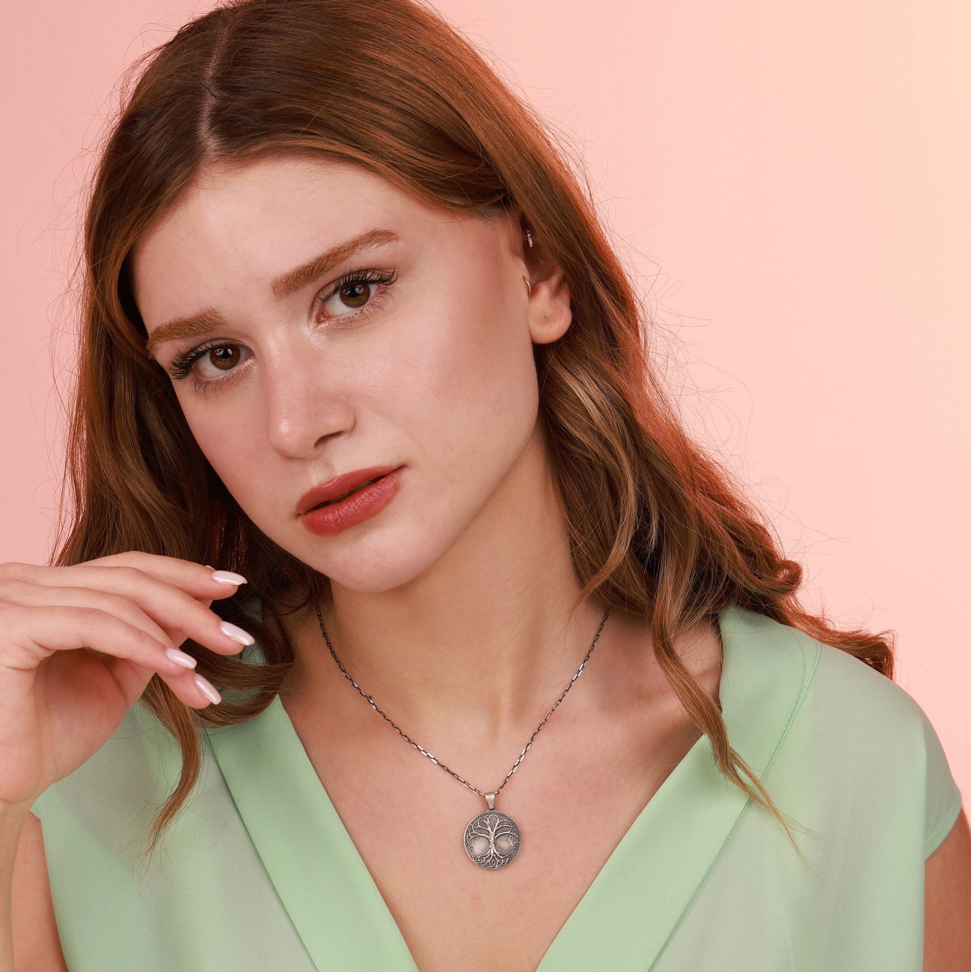 Woman wearing a green top and an oxidized necklace with a detailed tree of life pendant against a pink background