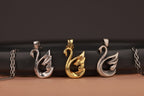 Three swan-shaped pendants in silver, gold, and bronze on a black and brown background.