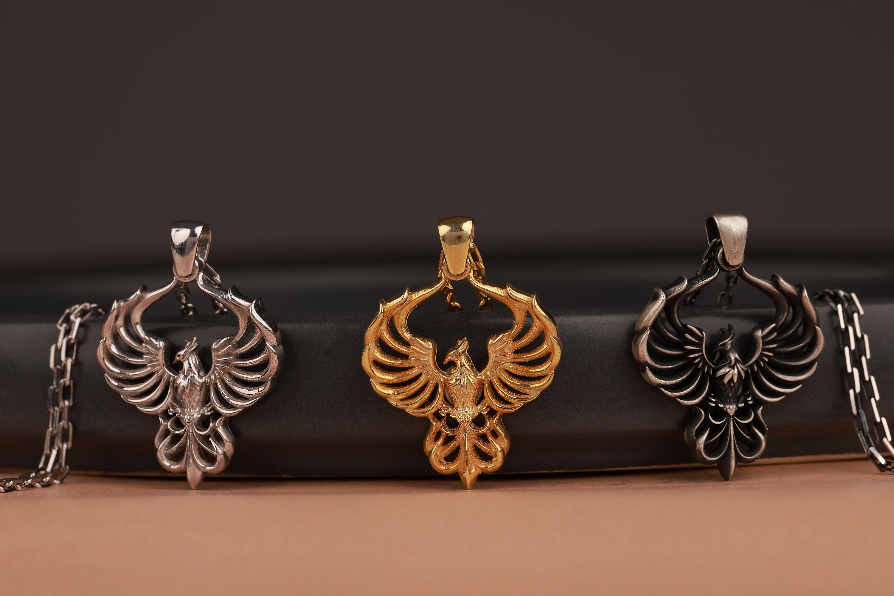 Three firebird-shaped pendants in silver, gold, and bronze on a dark surface.