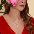 A woman wearing a gold necklace with a Hamsa hand pendant, holding pink flowers.