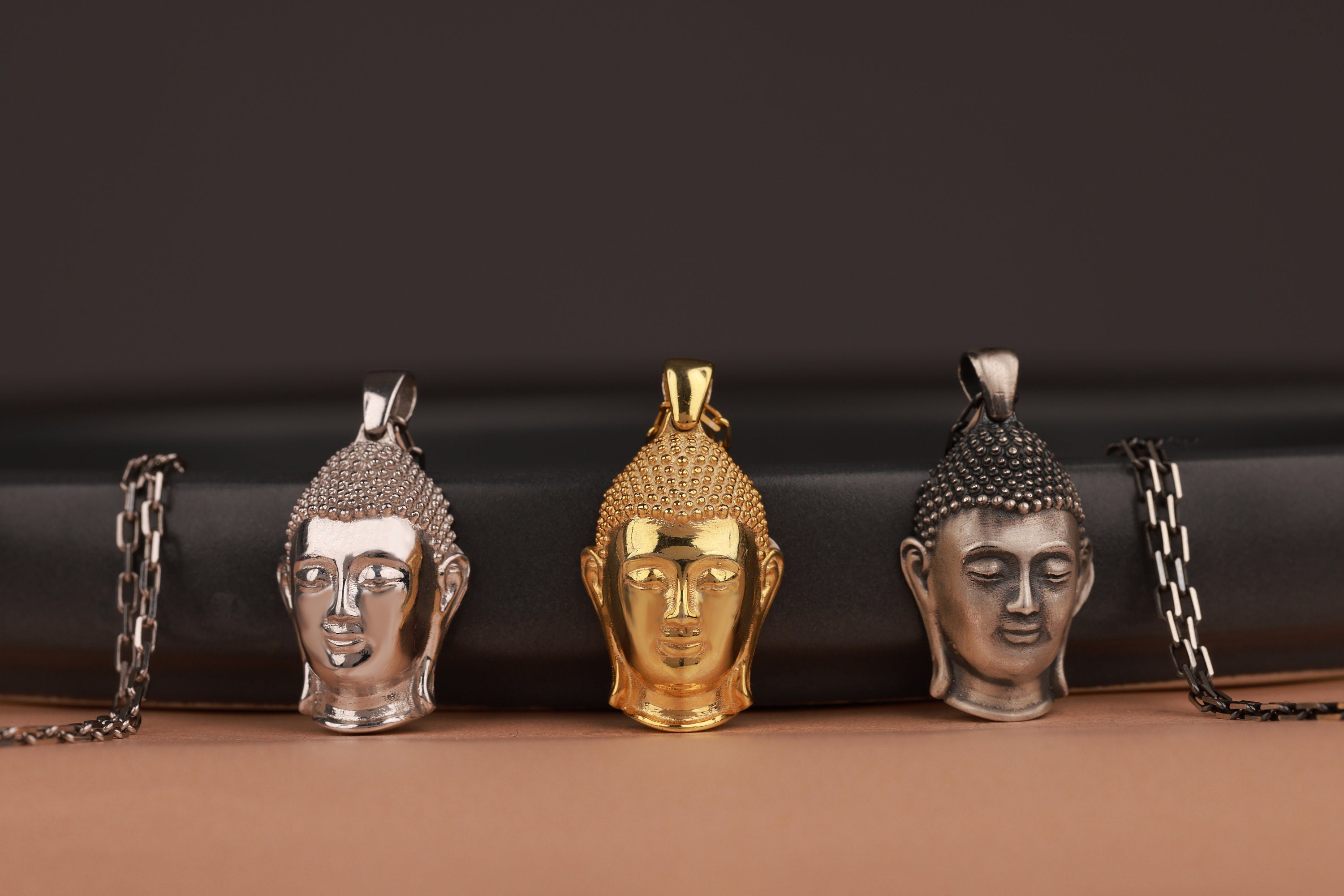 Three Buddha head pendants on chains against a dark background