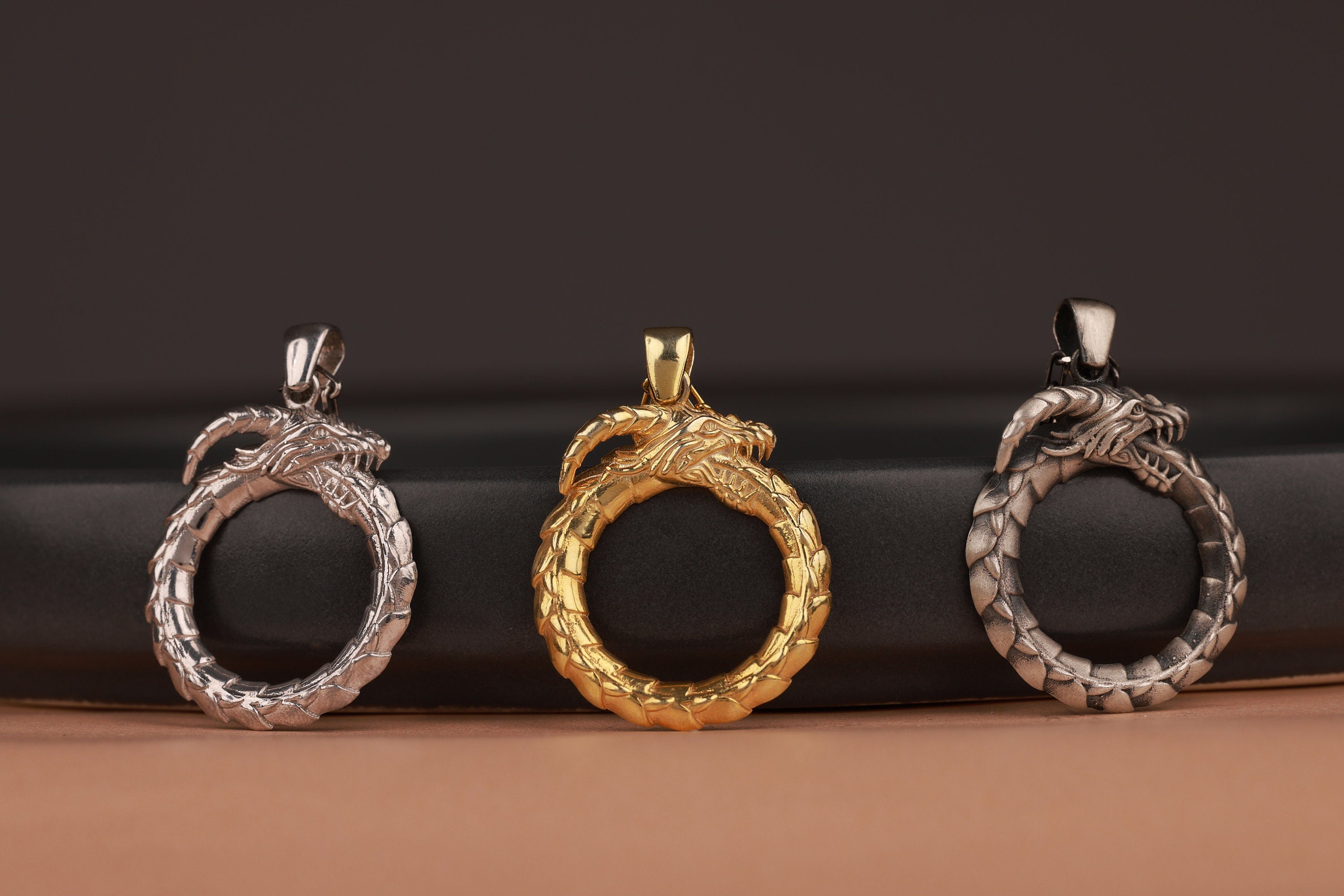 Three dragon-shaped pendants in silver, gold, and another metallic finish on a dark background.