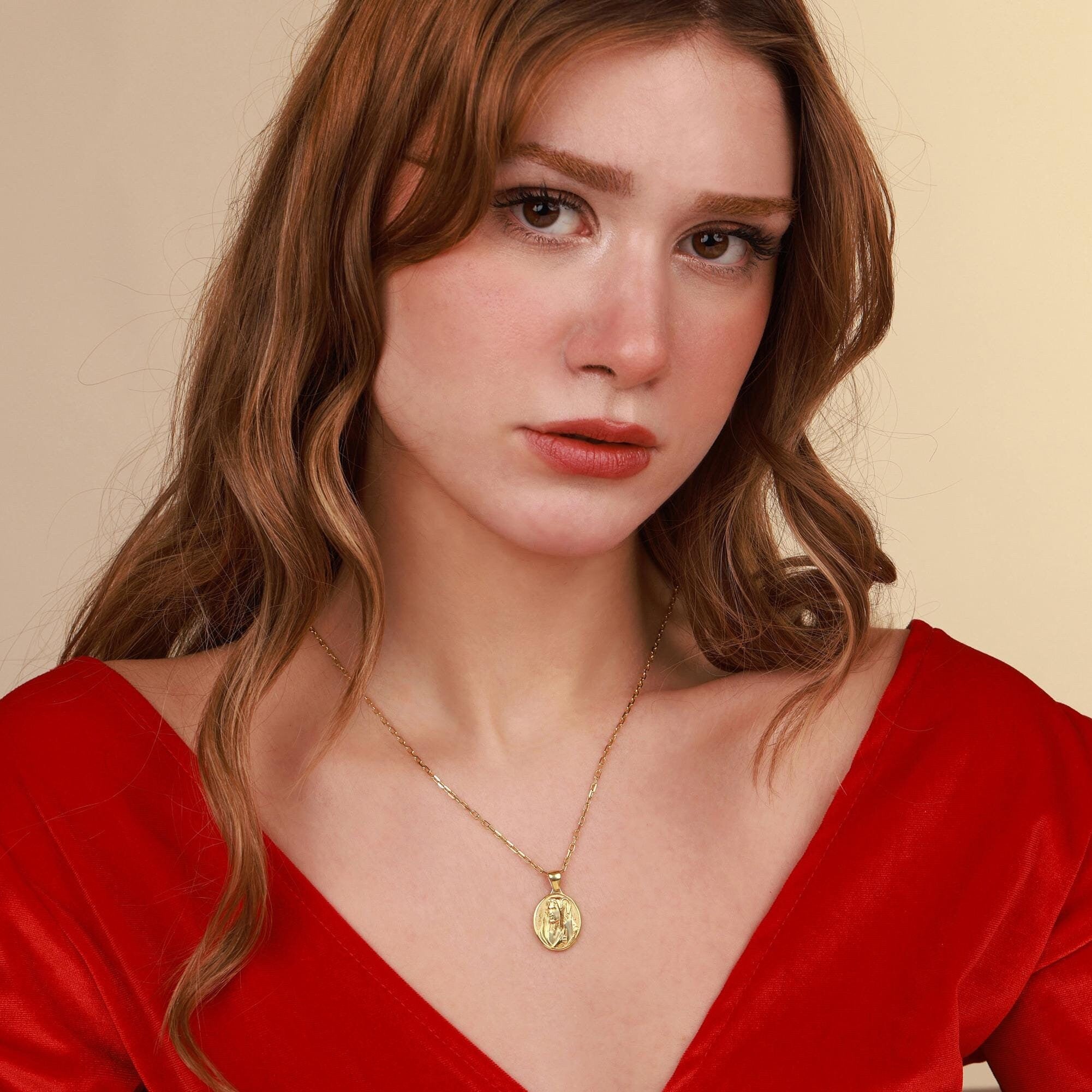 Woman wearing a gold jesus necklace with a red top against a beige background