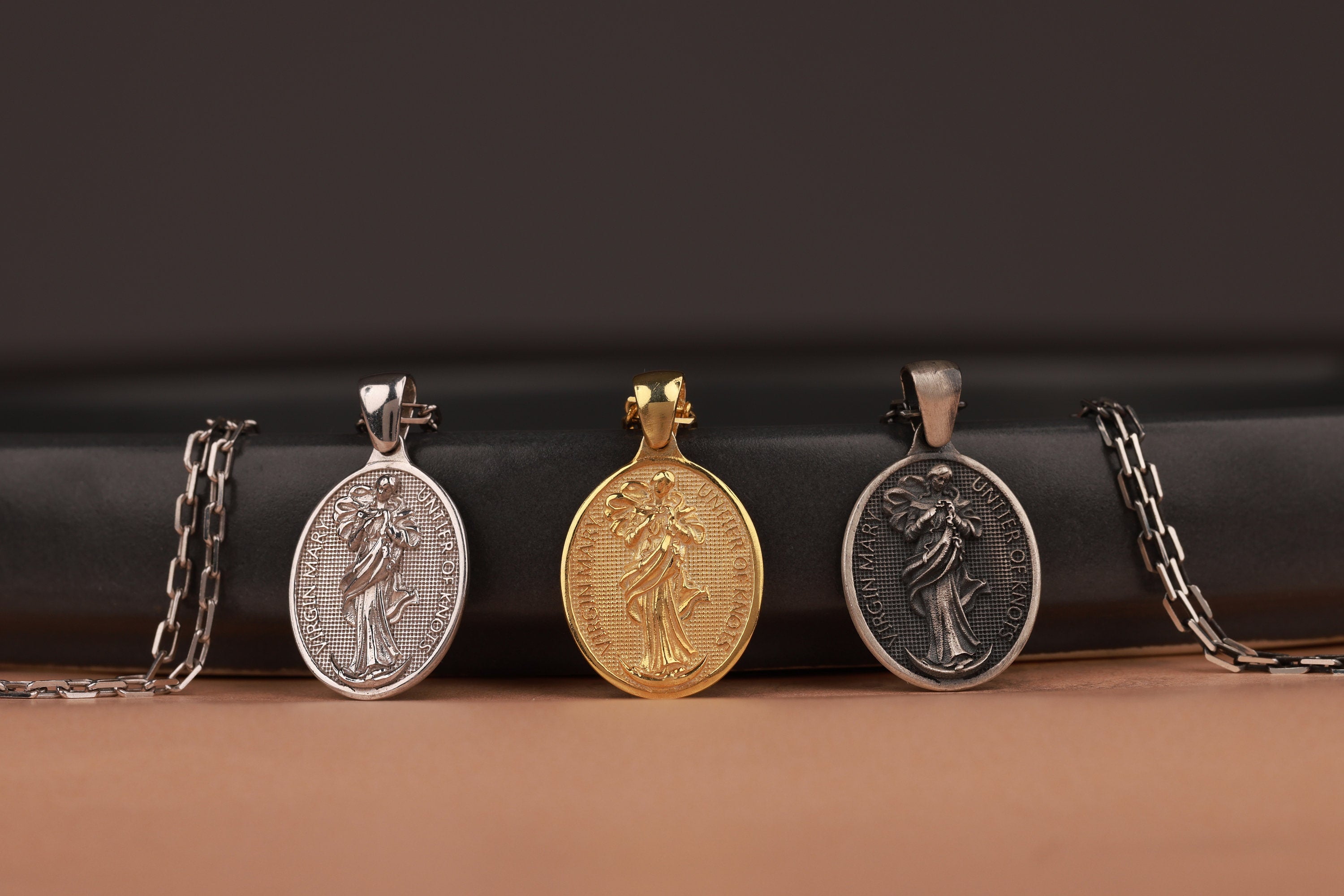 Three different Mary Undoer of Knots pendants on chains against a dark background