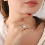 Close-up of a hand wearing a gold ring with a blue gemstone, touching the chin.