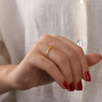 Hand wearing a gold ring with a pink gemstone against a neutral background