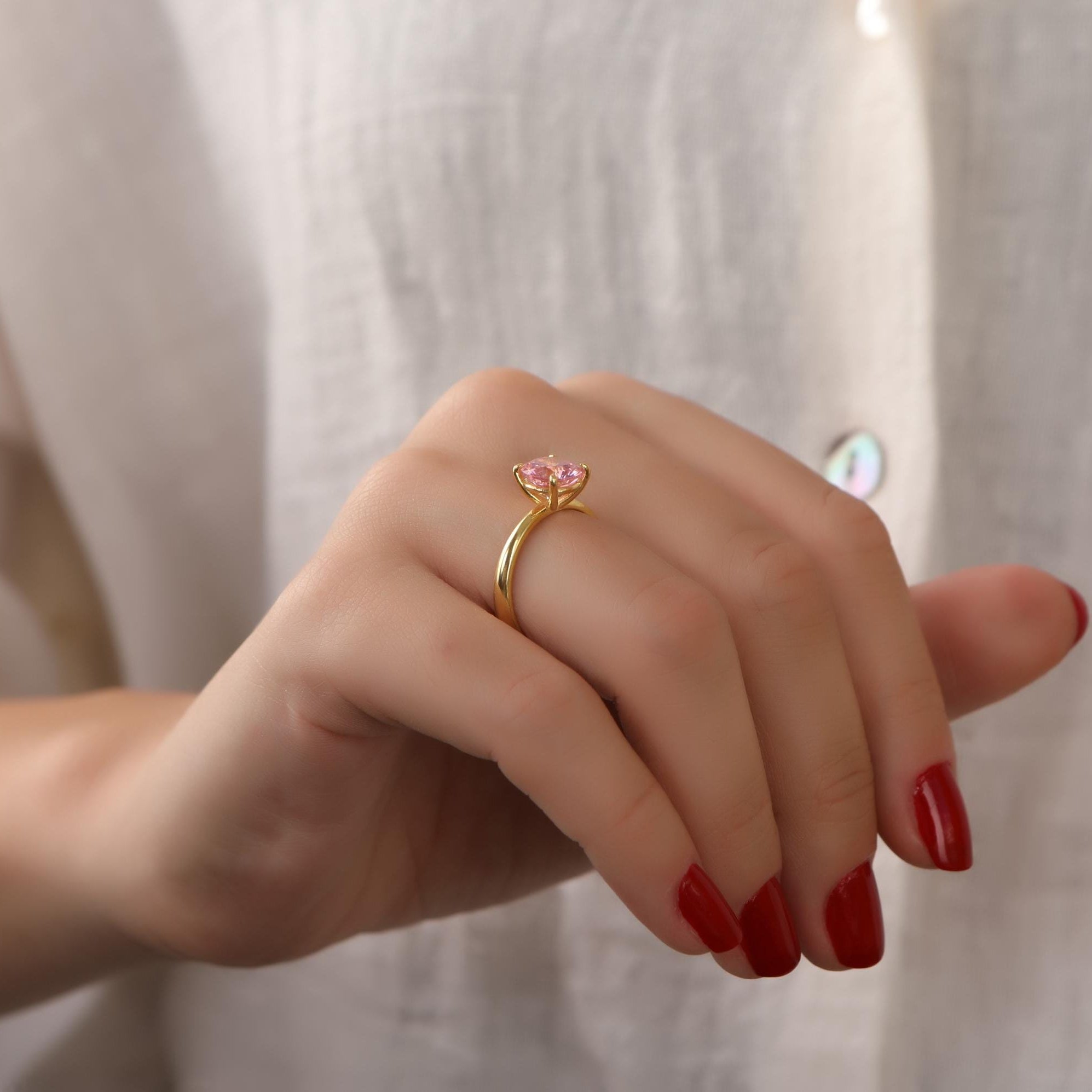 Hand wearing a gold ring with a pink gemstone against a neutral background