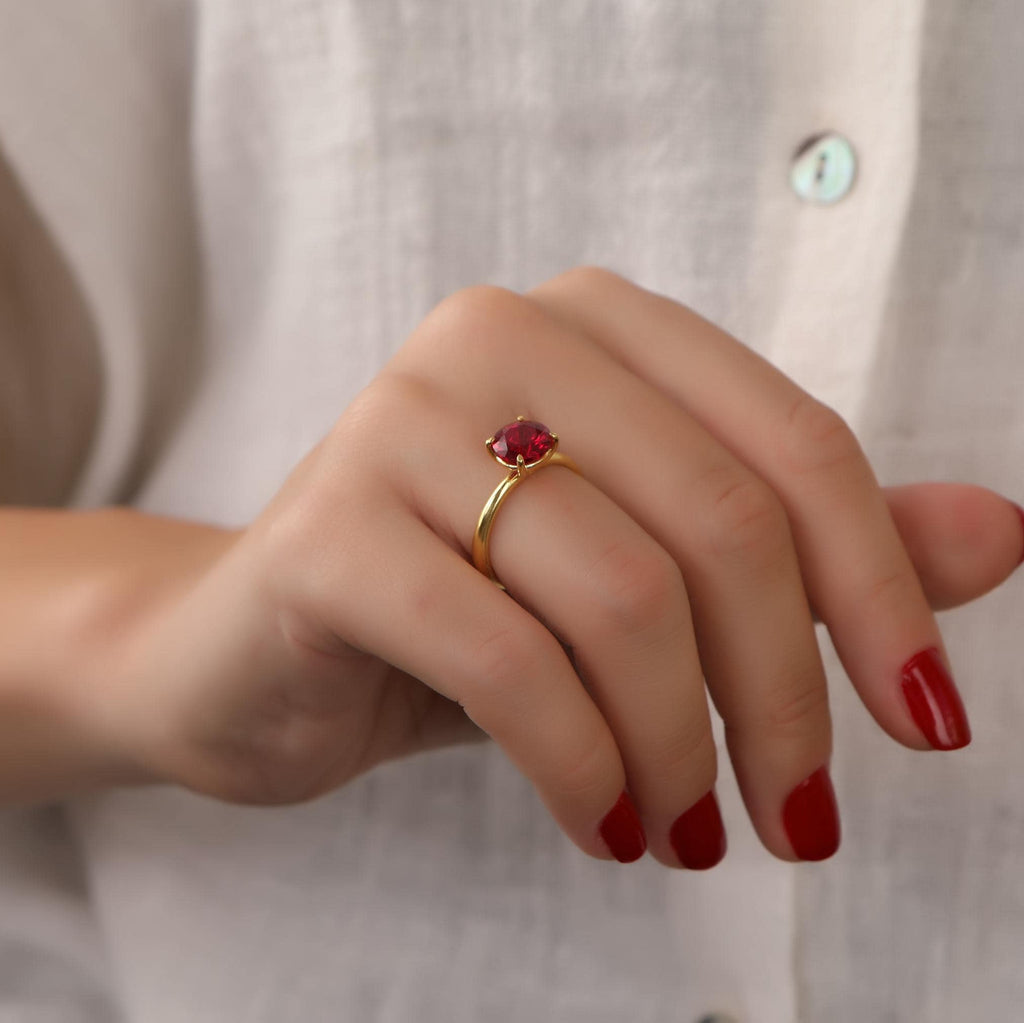 Close-up of a hand wearing a gold ring with a red gemstone, against a neutral background.