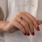 Close-up of a hand wearing a gold ring with a red gemstone, against a neutral background.