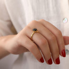 Close-up of a hand wearing a gold ring with a black stone, against a blurred white background.
