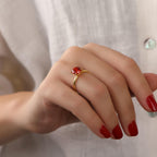 Hand wearing a gold ring with a red gemstone against a neutral background
