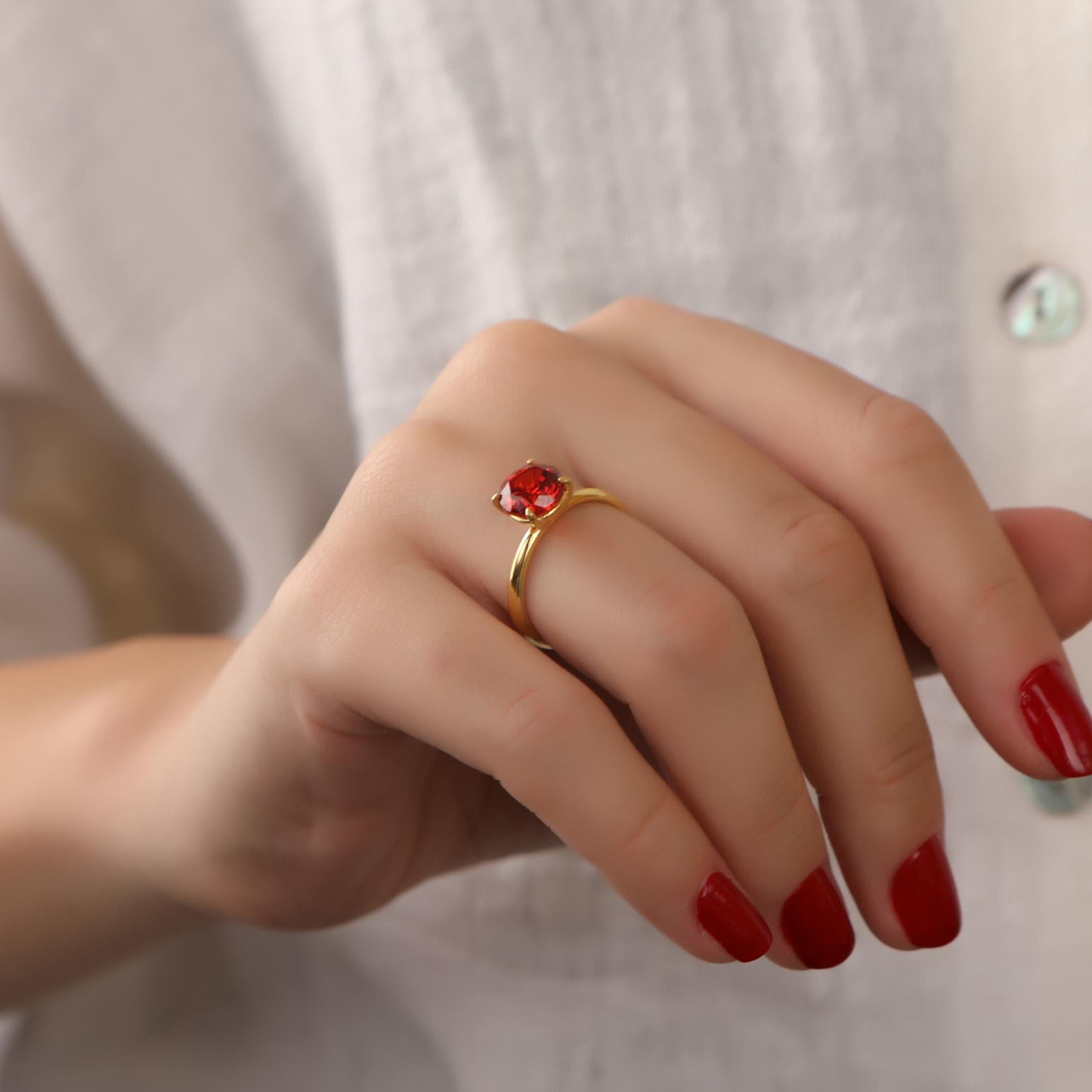 Hand wearing a gold ring with a red gemstone against a neutral background