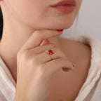 Close-up of a person wearing a gold ring with a red gemstone, holding their chin.