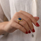 Hand wearing a gold ring with a blue gemstone against a white background